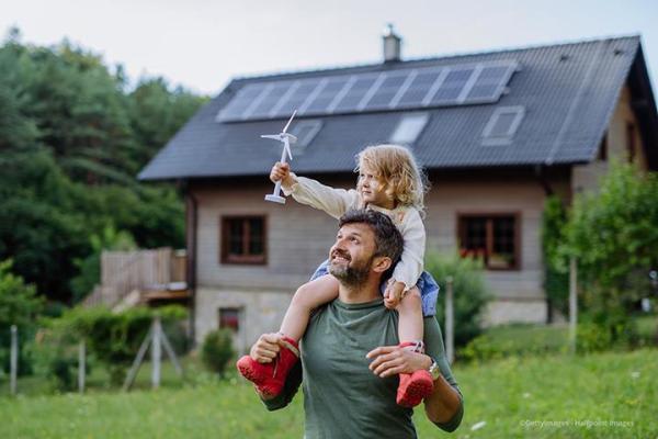 man with daughter sitting on his shoulders holding a mini wind turbine in front of house with solar panels